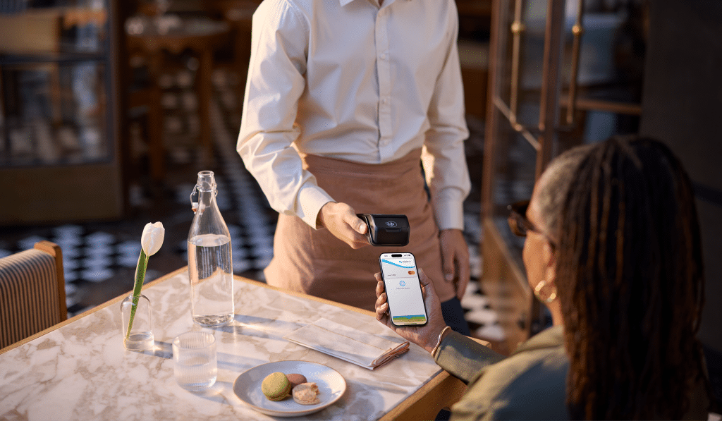 An employee pays for a business lunch using Apple Pay and SimpledCard on their iPhone at a payment terminal in a restaurant